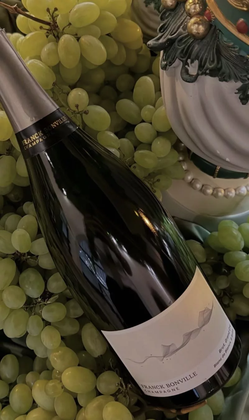 Franck Bonville Blanc de Blancs Grand Cru Champagne bottle displayed among fresh green grapes, highlighting the Chardonnay heritage of the Champagne house.