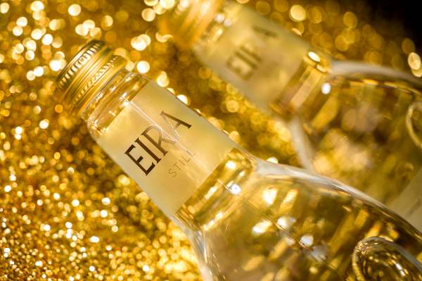 Close-up of an Eira Still Natural Mineral Water bottle with elegant gold branding against a sparkling golden background.