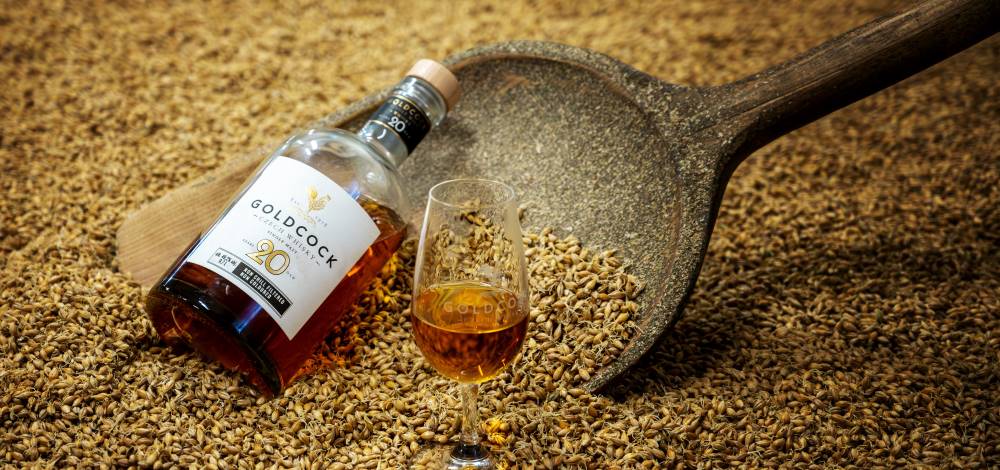 Gold Cock 20 Year Czech whisky bottle and tasting glass resting on malted barley, showcasing premium whisky ingredients.