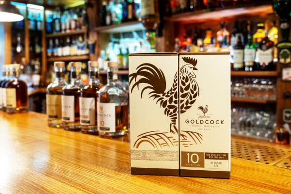 Gold Cock 10 Year Czech whisky gift box displayed on a bar counter with whisky bottles in the background.