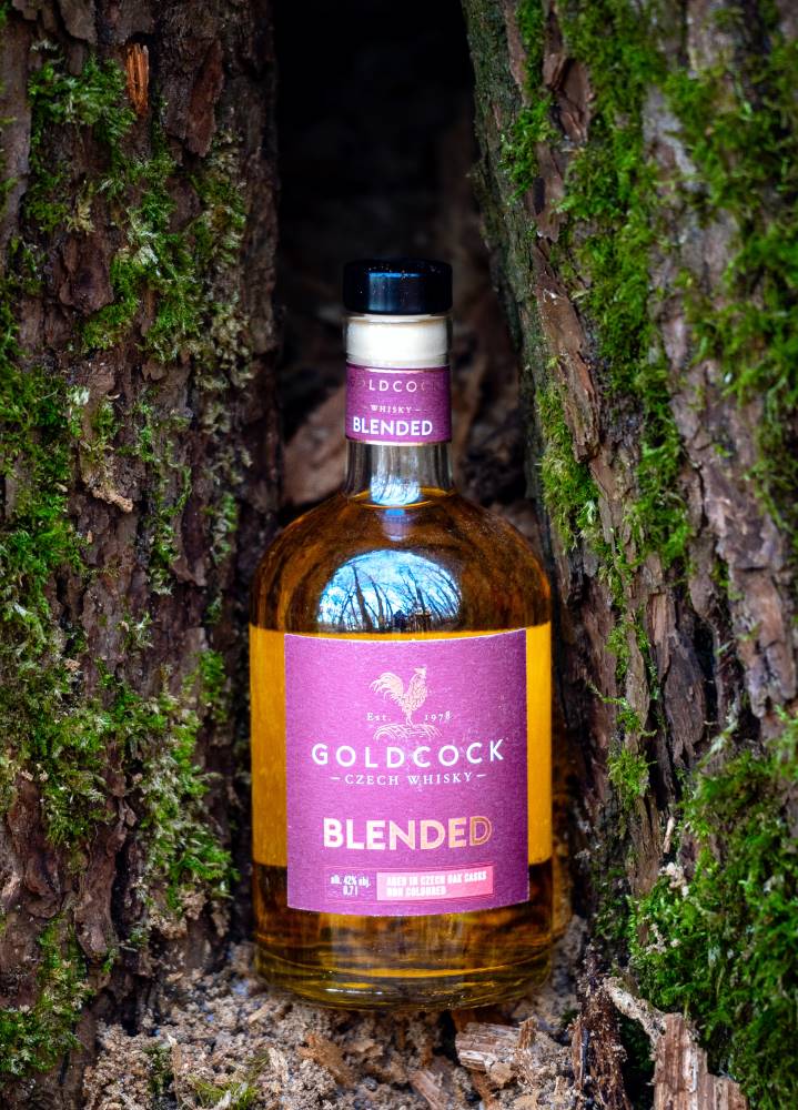 Gold Cock Blended Czech whisky bottle displayed between two moss-covered tree trunks in a natural forest setting.