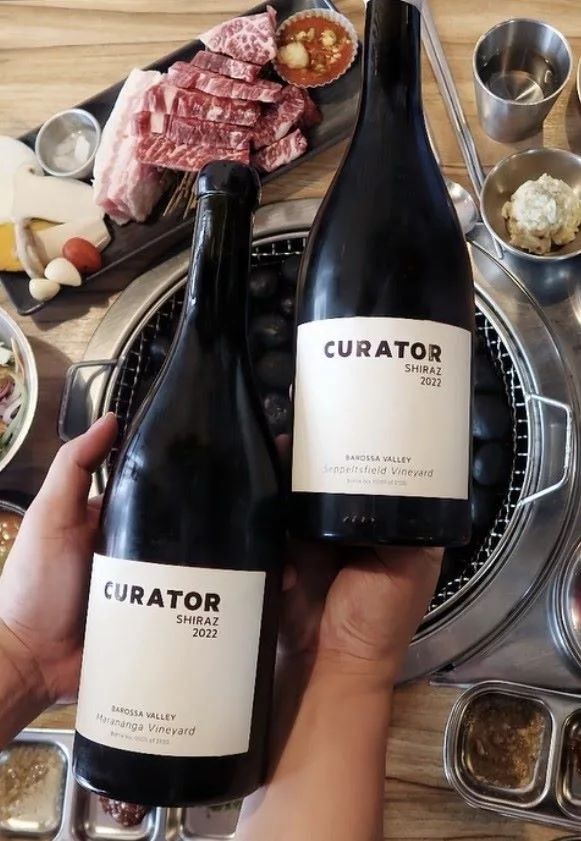 Two bottles of Curator Wine Co. Shiraz 2022 displayed over a Korean BBQ dining table, showcasing the wine as the perfect pairing for grilled meats.