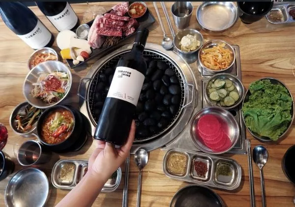 Curator Wine Co. red wine bottle held above a fully prepared Korean BBQ dining table, featuring premium Barossa Valley wine for shared dining experiences.