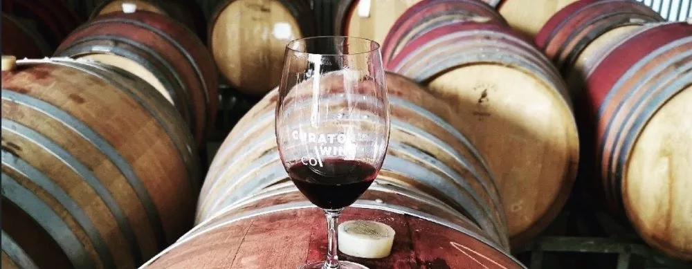 A glass of Curator Wine Co. red wine resting on an oak barrel inside the winery barrel room, surrounded by aging wine barrels.