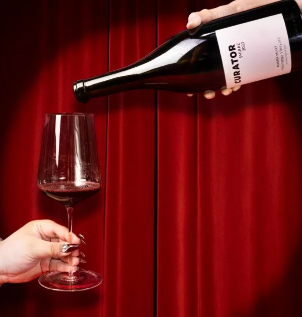 Curator Wine Co. Shiraz 2022 being poured into a wine glass against a rich red backdrop, highlighting the premium Barossa Valley red wine.